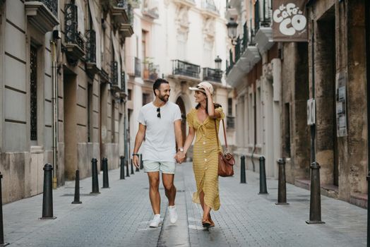 Stylish brunette girl in the hat and her boyfriend with a beard are walking together in the center of the old European street in Spain in the evening. A couple of young tourists enjoy Valencia.