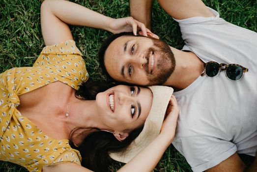 A smiling girl in a yellow dress and a hat is lying near her boyfriend with a beard on the grass in the park in Valencia. A couple of tourists is enjoying the summer in Spain.
