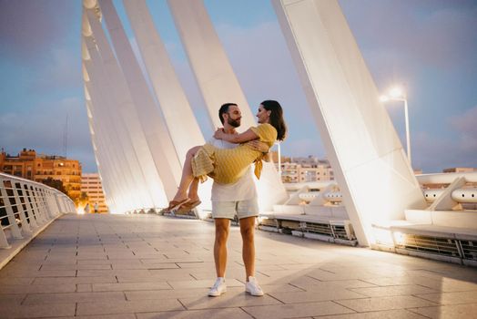 A man with a beard is holding his girl in a yellow dress in his arms on a white bridge in Valencia. A couple of tourists in full length on a date in the warm evening.