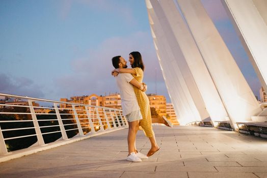 A man with a beard is hugging the waist of his girl in a yellow dress on a white bridge in Valencia. A couple of tourists in full length on a date in the warm evening.