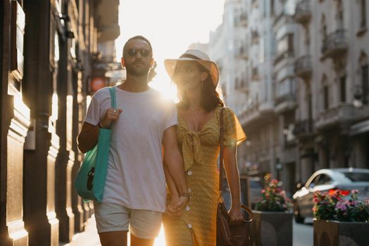 Girl in a hat and a yellow dress with a plunging neckline and her boyfriend with a beard and sunglasses are relaxing holding each other's hand in Spain. A couple of tourists on the sunset in Valencia.