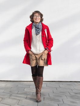 Curly woman in red duffle coat is standing by white wall with sunbeams. Smiling student. Modern lifestyle.