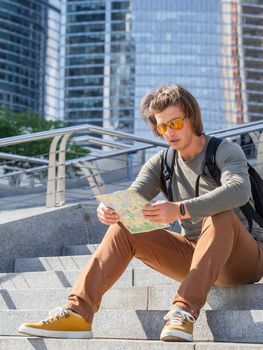 Young man sits on stone staircase in park and reads map. Solo-travelling around city. Urban tourism. Modern architectural landmarks.