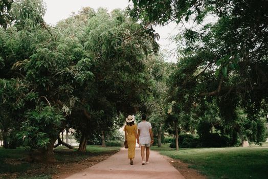 A photo from behind of a brunette girl in a yellow dress and her boyfriend who are walking on a sand path between trees in the Valencian park. A couple of tourists on a date in the warm evening.