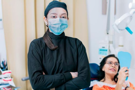 Dental doctor standing in clinic wearing mask, portrait of dentist crossing arms with patient in background, Modern Dental clinic. Dental procedures. portrait of a dentist woman with crossed arms