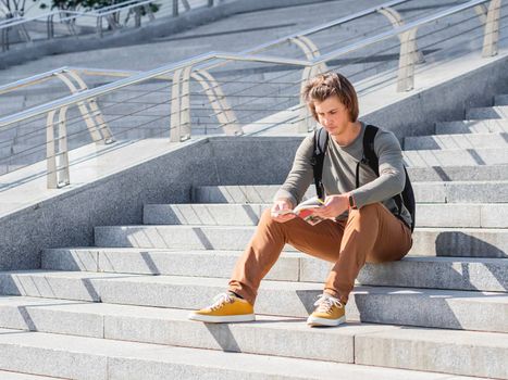 Young man sits on stone staircase in park and reads travel guide. Solo-travelling around city. Urban tourism. Modern architectural landmarks.