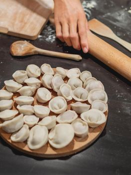 Woman makes pelmeni or dumplings - traditional dish of Russian cuisine made of dough and meat. Black kitchen table with flour and wooden utensils.