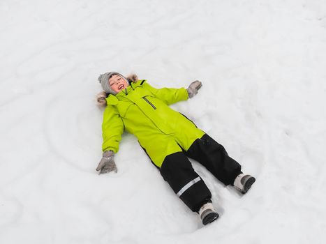 Laughing boy in green jumpsuit is making snow angel shape on snow. Joyful child playing outdoors in snowy weather. Top view on happy kid in colorful overall suit.
