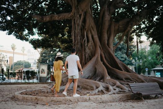 A young woman wears a hat and a yellow dress with her boyfriend go to an old Valencian Ficus Macrophylla tree in Spain. A couple of tourists are enjoying Valencia in the evening.