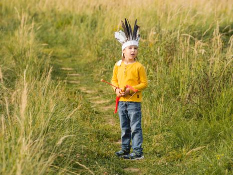 Little boy is playing American Indian on field. Kid has handmade headdress made of feathers and bow with arrows. Costume role play. Outdoor leisure activity. Fall season.