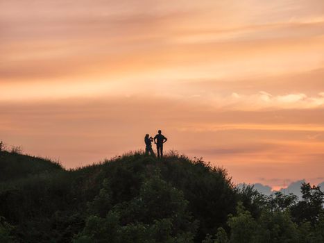 Silhouette of couple meeting sunset on hill. Woman takes picture of clouds on gorgeous orange sky background. Romantic evening outdoors.