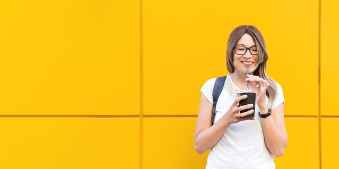 Wide smiling woman drinks coffee with black straw. Portrait of happy female with tasty cappuccino on monochrome yellow wall background. Bright and colorful backdrop. Student with coffee to go.