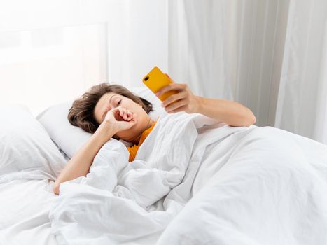 Sleepy woman is lying in bed, completely covered with white blanket. Smartphone used as alarm clock. It's hard to wake up early in morning. Woman does not get enough sleep.
