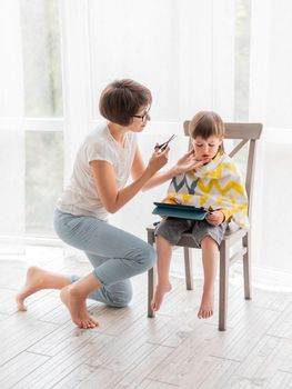 Mother cuts her son's hair by herself. Little boy sits, covered with cloth, and holds digital tablet. New normal in case of coronavirus COVID-19 quarantine and lockdown.