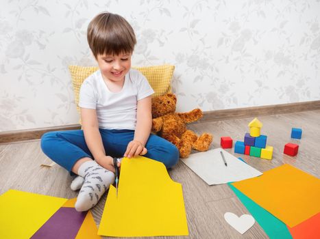 Toddler boy learns to cut colored paper with scissors. Kid sits on floor in kids room with toy blocks and teddy bear. Educational classes for children. Developing feeling sensations and fine motor skills at home.