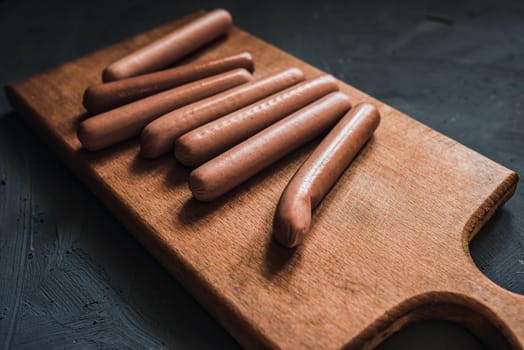 Boiled fried sausages sausages lie on a wooden kitchen board scratched against a dark concrete background.