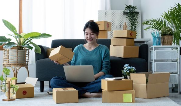 Happy young asian woman startup small business freelance holding parcel box and computer laptop and sitting on floor, Online marketing packing box delivery concept.