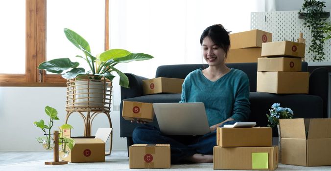 Happy young asian woman startup small business freelance holding parcel box and computer laptop and sitting on floor, Online marketing packing box delivery concept.