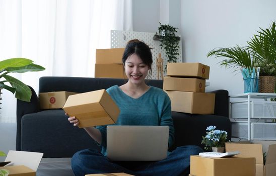 Happy young asian woman startup small business freelance holding parcel box and computer laptop and sitting on floor, Online marketing packing box delivery concept.