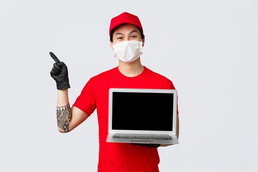 Smiling asian delivery man in uniform, red cap and t-shirt, wearing protective mask and gloves to prevent covid 19 spread, deliver online orders during self-quarantine isolation, invite visit webpage.
