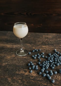 A transparent glass with milk. Blue fresh blueberries are scattered on an old brown wooden cracked table.