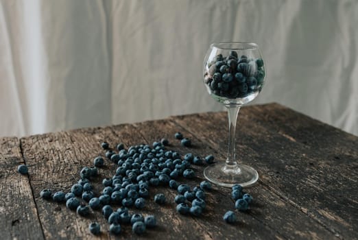 Fresh blue matte round blueberries filled in a transparent glass stands on a wooden brown shabby textured table and berries are scattered everywhere