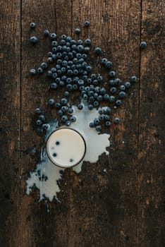 Berries are thrown into the transparent glass making splashes of milk. Blue fresh blueberries are scattered on an old brown wooden cracked table. White water pour down on the table.