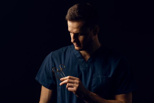 Handsome doctor with surgical scissors on dark background. Confident man holding medical equipment in hands and smiling. Happy male posing in studio