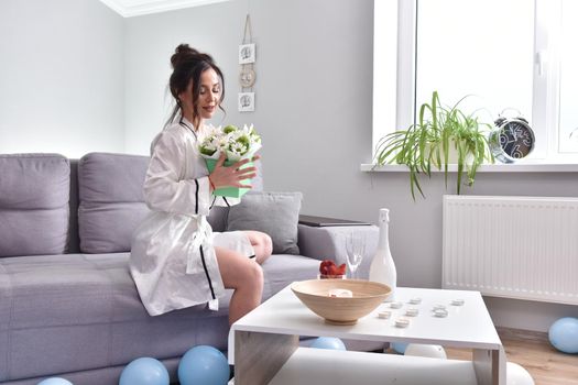 Beautiful brunette woman dressing gown with bouquet indoors. Female portrait in living room