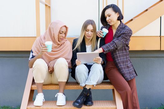 Trendy young multiracial girls students in casual clothes drinking takeaway coffee and using tablet while sitting on stairs in city and preparing for exams