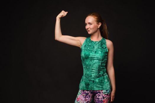 Front view of strong red haired woman in green uniform looking at camera and posing on black isolated background. Muscular girl keeping fit and training. Concept of health and workout.