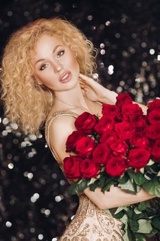 Portrait of gorgeous young blonde woman with curly hair and nude make-up in expensive elegant dress looking at camera holding beautiful red roses. Sparkling background. Bokeh.