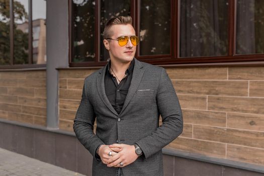 Stylish young guy portrait businessman of European appearance in a gray jacket and black shirt in sunglasses on the street outdoor.