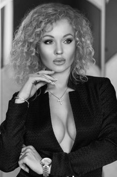 Black and white portrait of gorgeous blonde woman with curly hair in expensive formal jacket dressed up on ideal naked breast. She is stunning and sexy. Looking away with thoughtful look. Wearing expensive watch, bracelet and pending in shape of religious cross.