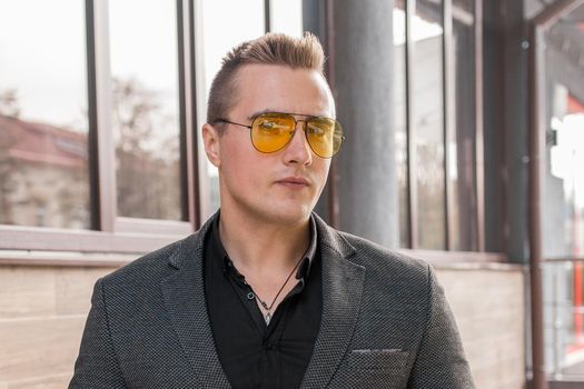 Stylish young guy portrait businessman of European appearance in a gray jacket and black shirt in sunglasses on the street outdoor.