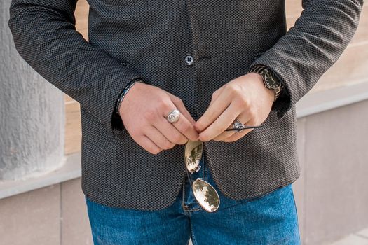 A man in a gray jacket and blue jeans holds sunglasses in his hands, close-up.