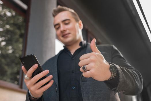 A young positive man in a grey jacket and black shirt holds a mobile phone or smartphone and points the class with his finger up on the street outdoor, close-up.