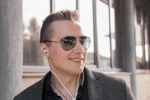Portrait of a smiling young guy of European appearance, an attractive businessman in sunglasses, listening to music in headphones on the street outdoor.