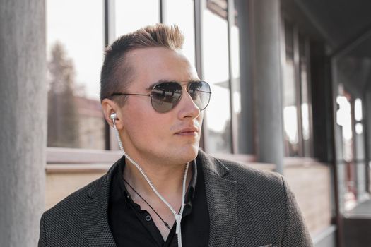 Portrait of a serious young guy of European appearance, an attractive businessman in sunglasses, listening to music in headphones on the street outdoor.