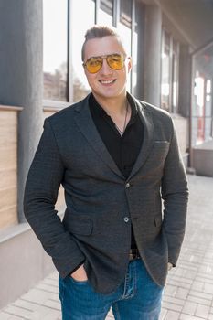 Stylish positive young attractive guy of European appearance businessman portrait in jacket, shirt and jeans, in sunglasses on the street outdoor.