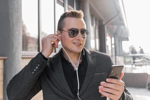 Portrait of a smiling stylish guy of Caucasian appearance of a businessman in sunglasses, jacket and shirt, adjusts his headphones with his hand listening to music from a mobile phone on the street outdoor.