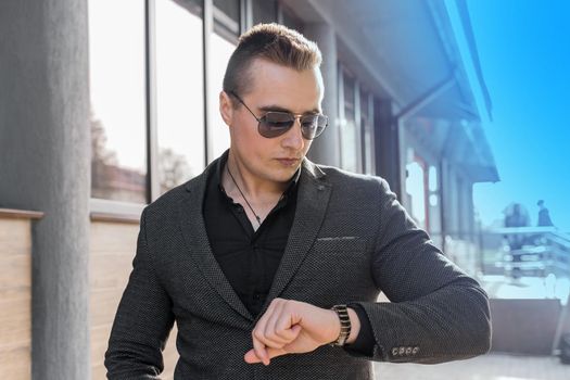 A young stylish, businessman of European appearance in sunglasses and a jacket with a shirt looks at the time on a hand watch on the street outdoor.