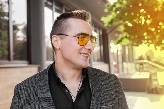 Stylish young smiling guy businessman of Caucasian appearance portrait in sunglasses, jacket and shirt on the street outdoor.