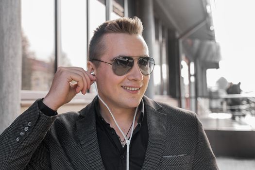 Portrait of a smiling young guy of European appearance, an attractive businessman in sunglasses, listening to music in headphones on the street outdoor.