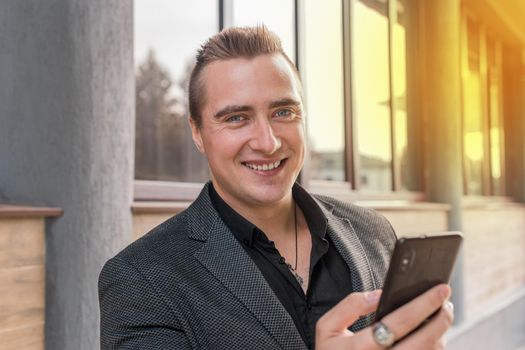 Portrait of a smiling, cheerful man of Caucasian appearance of a businessman in a jacket and shirt with a mobile phone in his hands on the street outdoor.