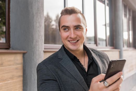 Portrait of a smiling, cheerful man of Caucasian appearance of a businessman in a jacket and shirt with a mobile phone in his hands on the street outdoor.