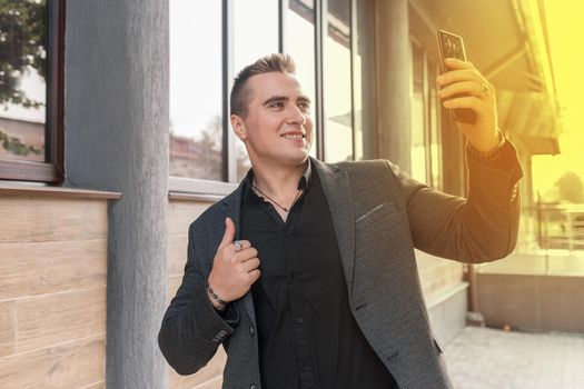 Businessman smiling man portrait of beautiful European appearance, in a jacket and shirt makes a selfie or talks on a video call on a smartphone on the street outdoor.