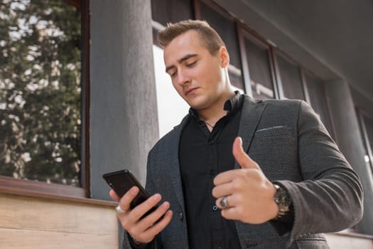 A portrait of a stylish young guy of Caucasian appearance in a jacket and shirt uses a smartphone and shows the class with a finger up on the outdoor street. close-up.