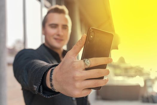 A young satisfied guy holds a smartphone or mobile phone in his hand, takes a selfie or talks on video on the outdoor street, close-up.