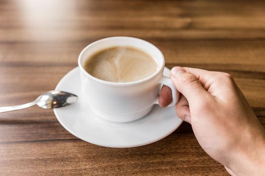 A man takes a cup of coffee on a saucer with his hand against the background of a wooden table in a cafe.